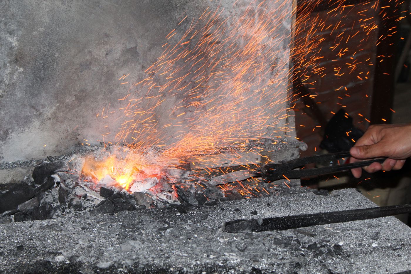 A coal forge burning bright orange with sparks rising and a steel bar in the fire