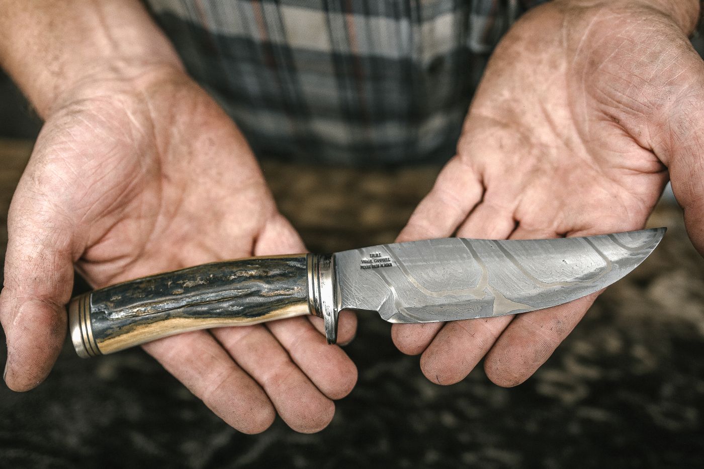 A finished hunting knife with a stag horn handle held in two weathered hands