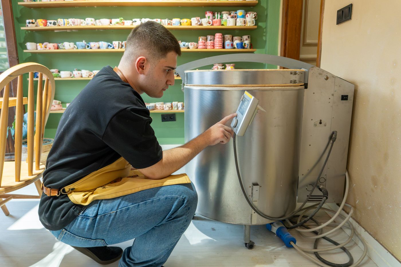 A potter crouched at a top-loading electric kiln operating a digital control panel