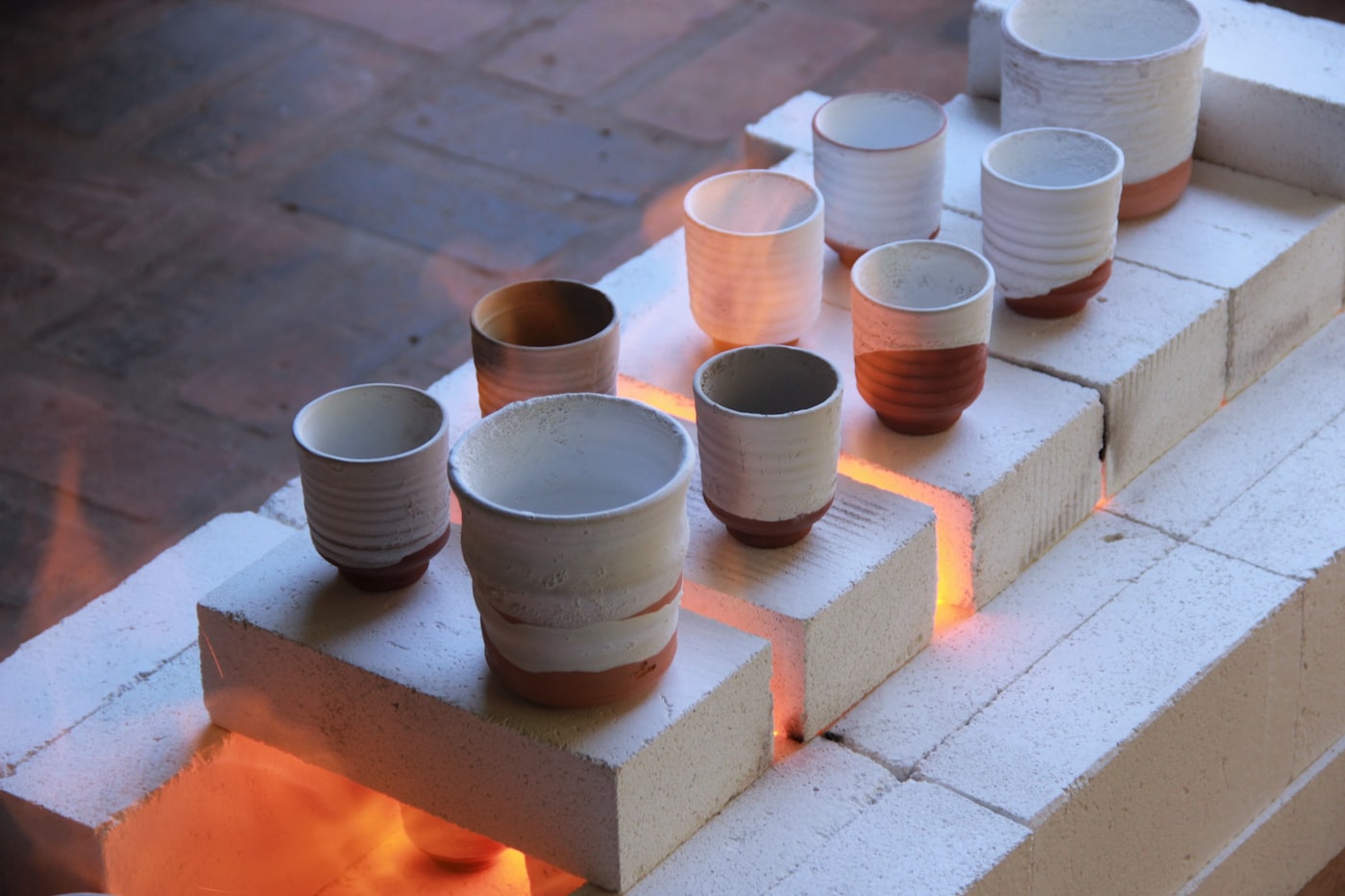 Glazed ceramic cups arranged on kiln bricks with the orange glow of a firing behind them