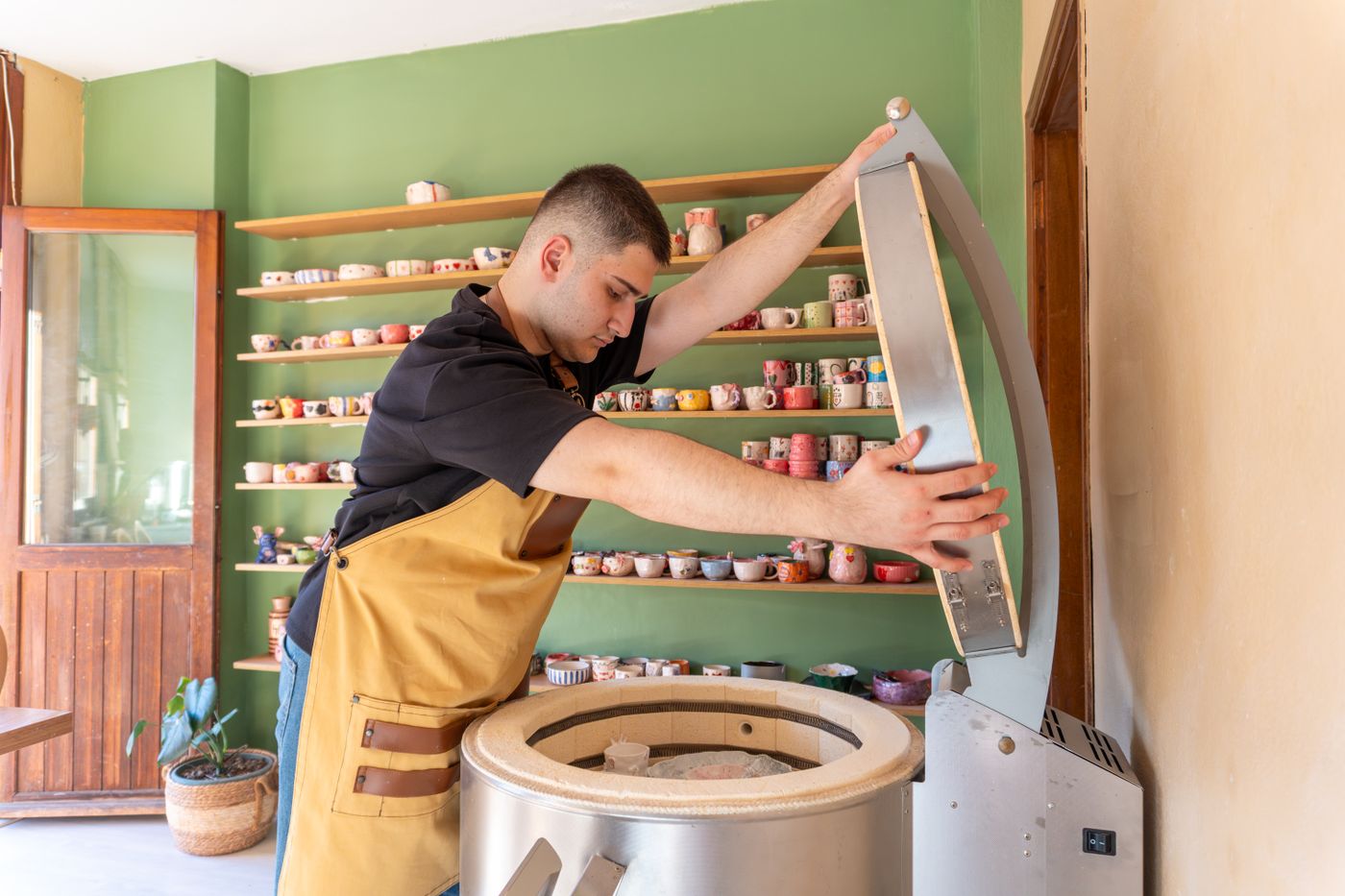 A potter lifting the hinged lid of a top-loading electric kiln in a studio lined with glazed work