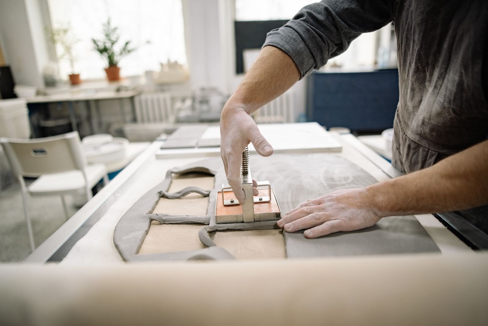 A potter pressing a tool into a flat clay tile on a work table