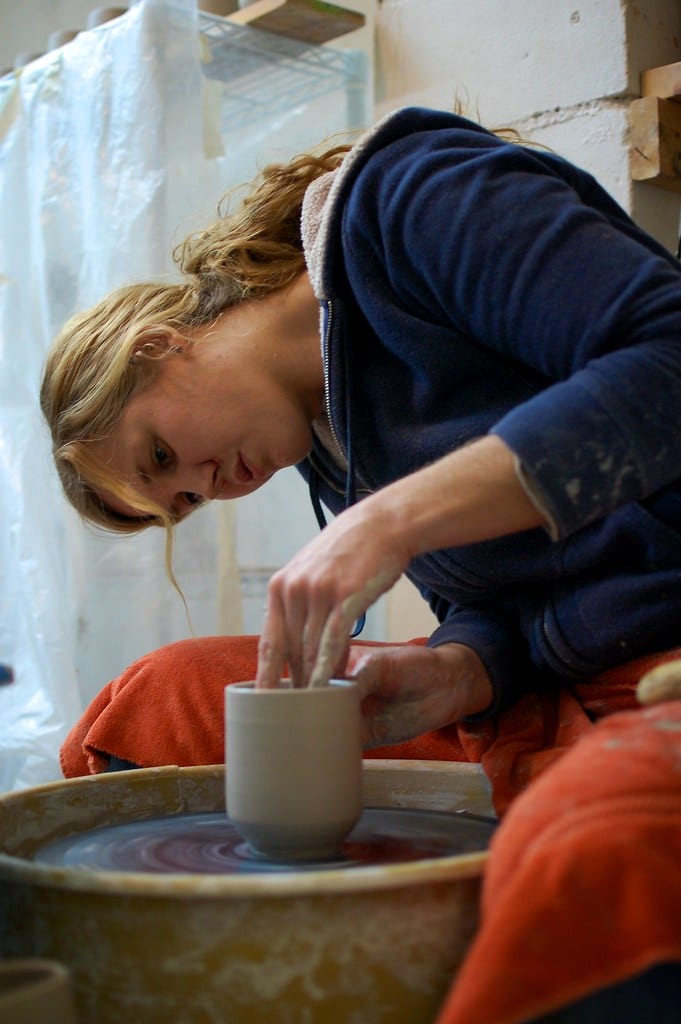 A potter's hands opening a centered mound of clay on a wheel head, clay slurry visible on the wheel and splashpan, studio setting