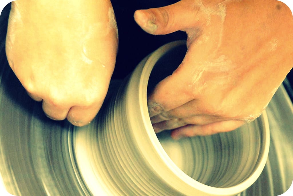 Close-up of hands shaping the walls of a clay cylinder on a pottery wheel, clay slurry covering the hands and lower walls