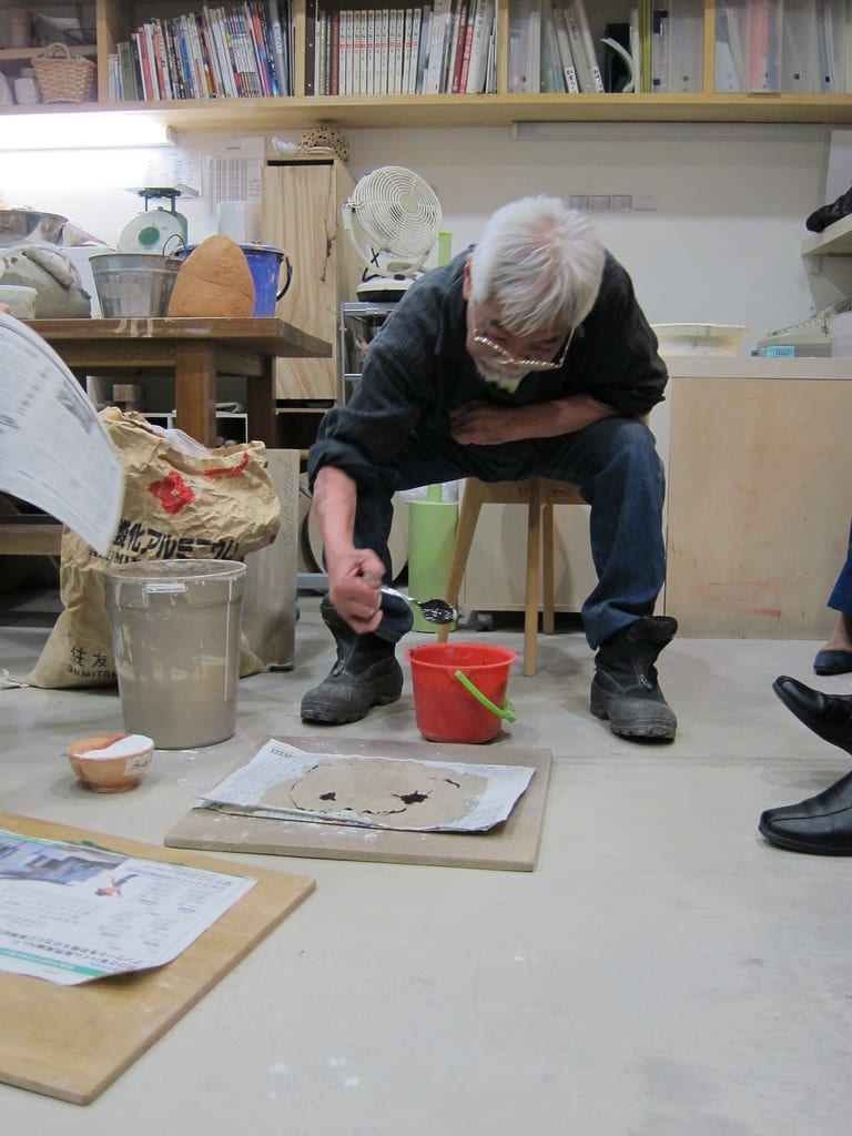A master potter demonstrating at a wheel with an audience watching, in a well-lit workshop studio setting with shelves of tools and ceramics in background