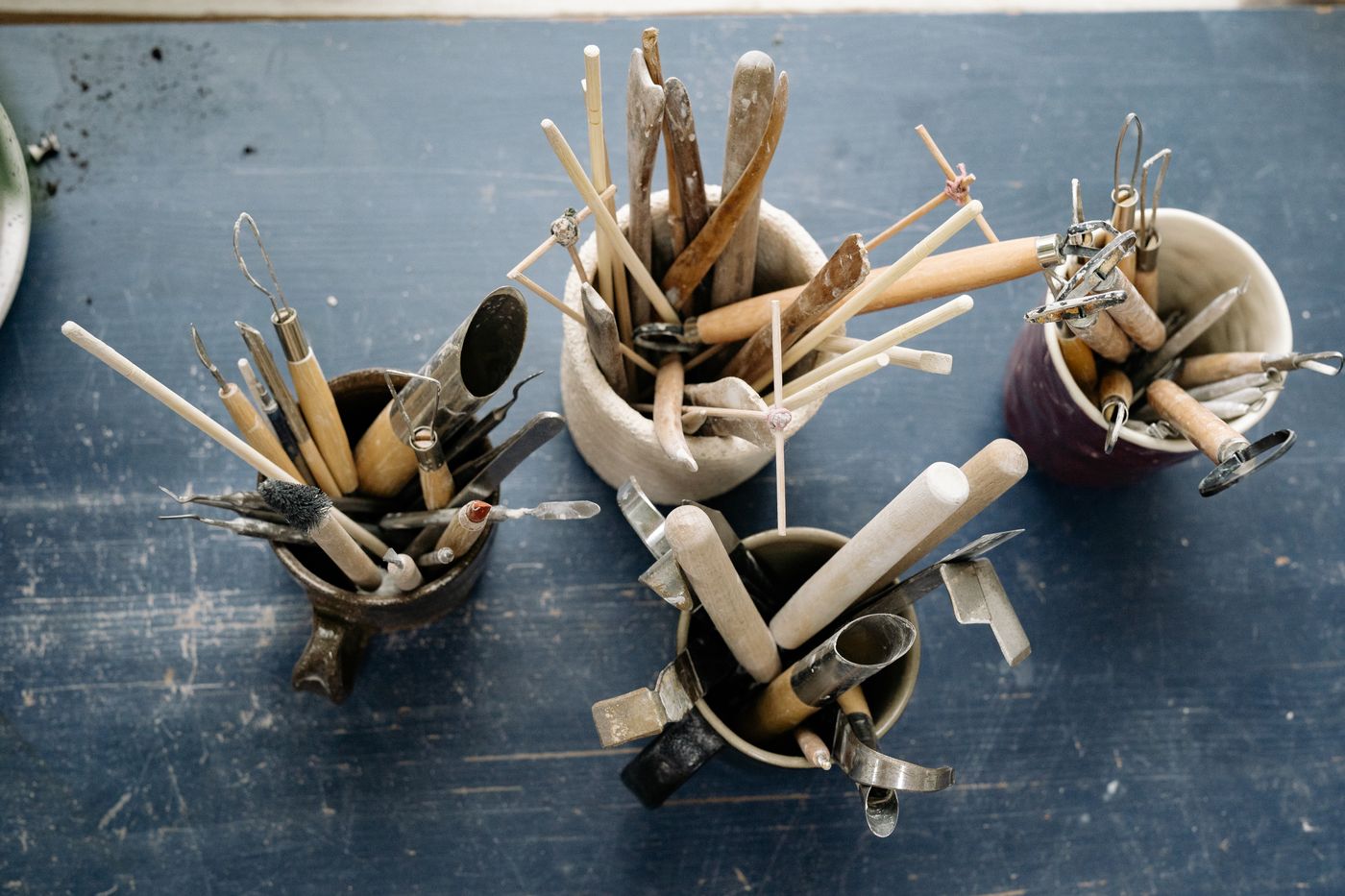 Wooden and wire pottery tools standing upright in thrown mugs on a dark workbench