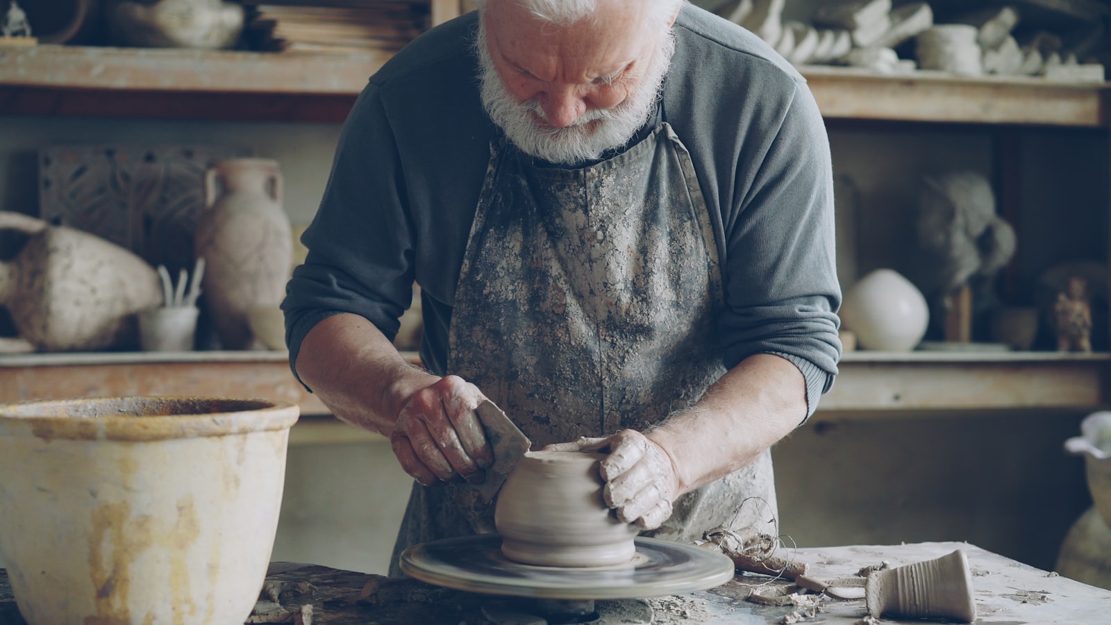 A potter seated at a full-size floor pottery wheel, shaping the lower wall of a tall clay vessel