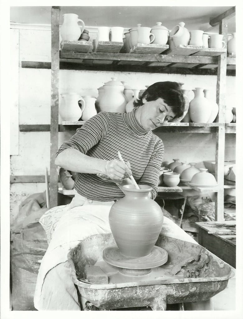 A potter throwing a jar at a full-size electric wheel surrounded by studio shelves
