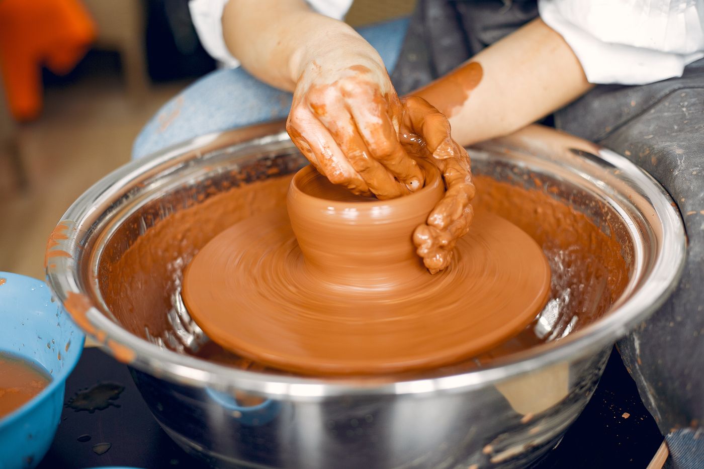 Hands centering a mound of terracotta clay on a full-size pottery wheel with a metal splash pan