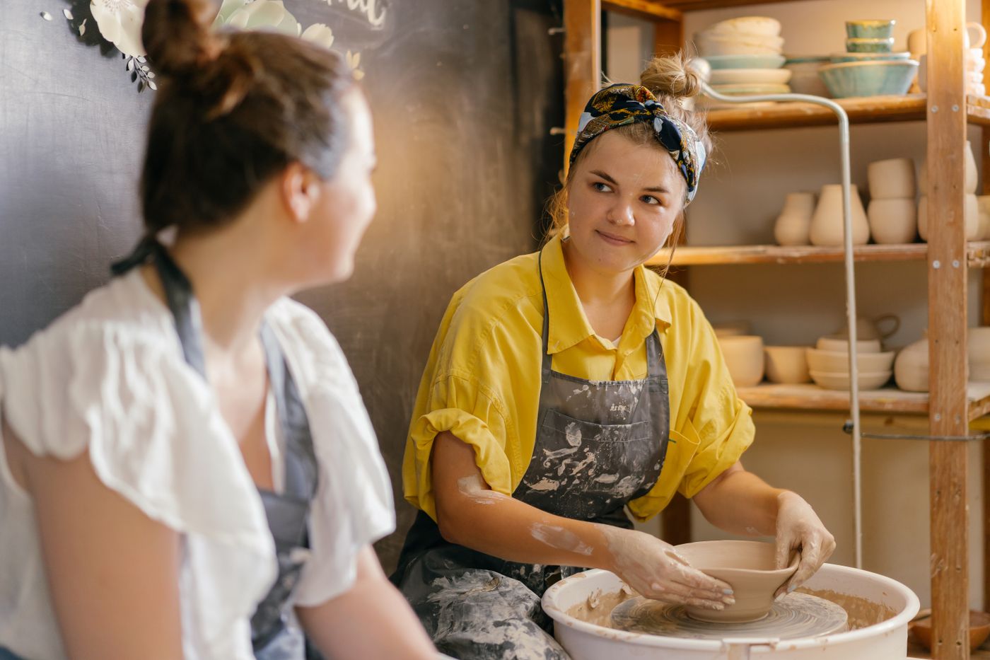 Two potters working together at a wheel in a studio lined with shelves of drying ware