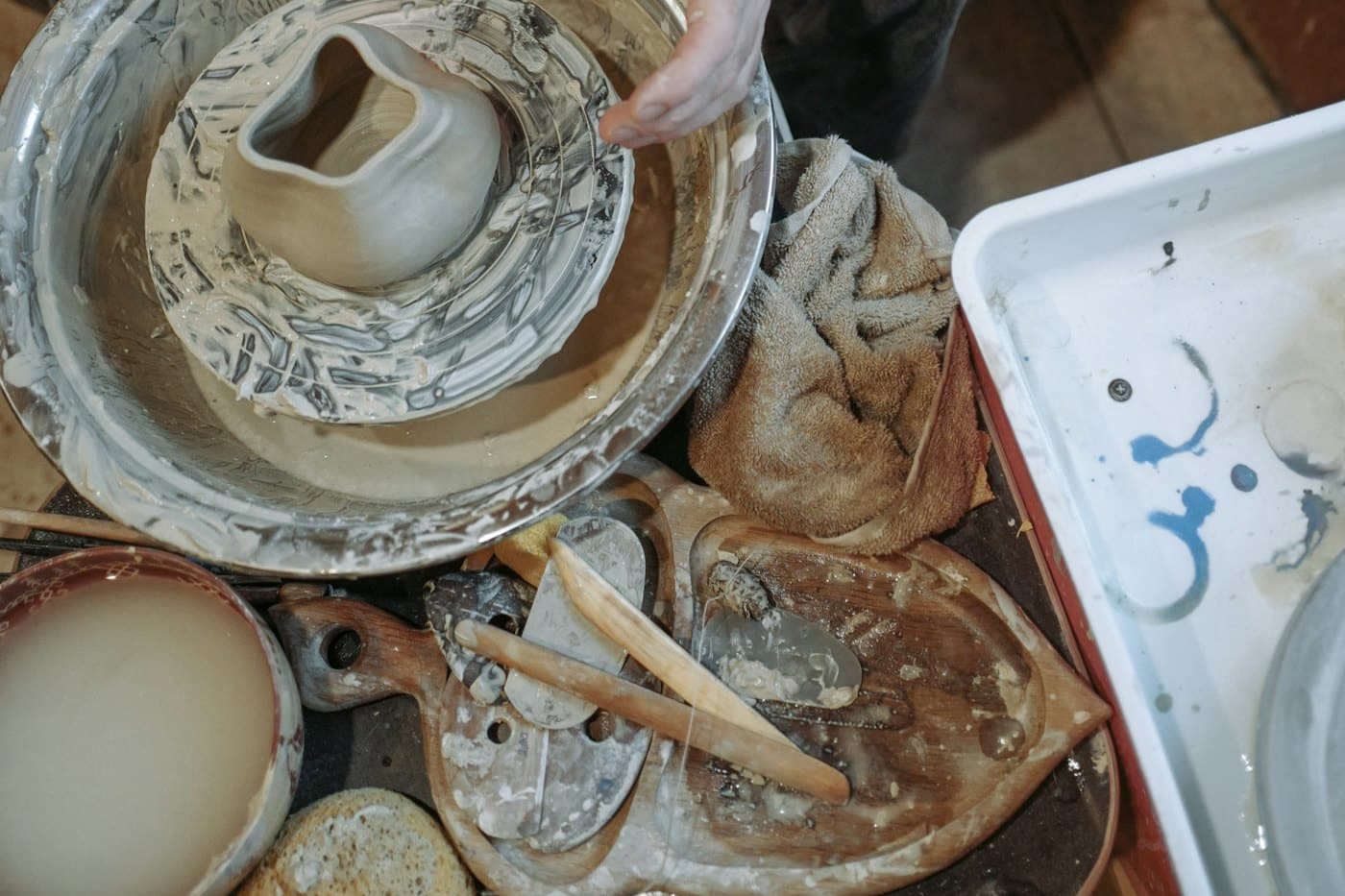 A small thrown pot standing on a clay-covered wheel head inside a splash pan, with tools alongside